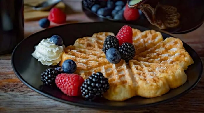 Healthy breakfast waffle topped with mixed fresh berries, whipped cream, and maple syrup being poured on a dark plate.
