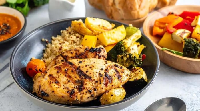 Healthy meal prep bowl with a grilled whole chicken breast, quinoa, roasted potatoes, broccoli, and grilled vegetables in a dark ceramic bowl.