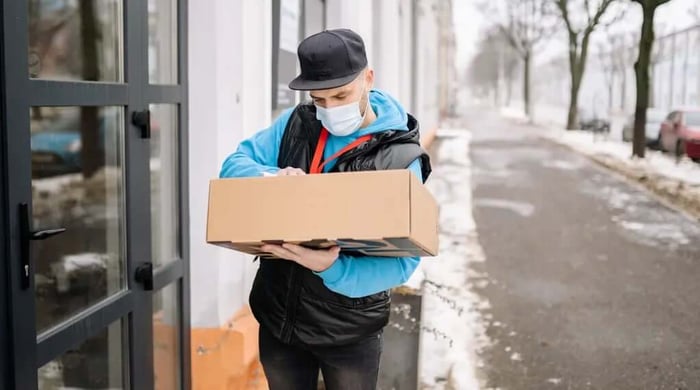 Healthy meal delivery courier in a face mask signing a clipboard while holding a cardboard package outside a building on a snowy street.