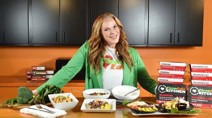 Healthy meal prep recipes showcased by woman in kitchen with plated meals and stacked Clean Eatz boxes.