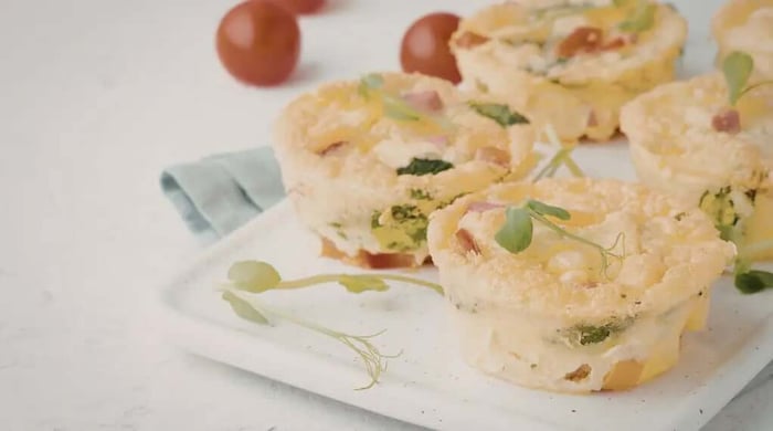 High protein breakfast meal prep egg cups filled with ham, spinach, and melted cheddar garnished with microgreens on a white cutting board beside cherry tomatoes.
