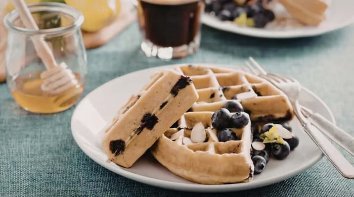 High protein food served as blueberry waffles with sliced almonds and fresh blueberries on a white plate alongside honey and black coffee.