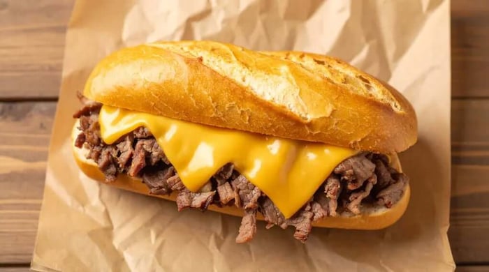 High protein food served as a Philly cheesesteak with shaved beef and melted yellow American cheese sauce on a toasted crusty hoagie roll over parchment paper.