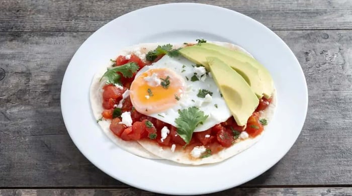 High protein food served as huevos rancheros with a fried egg over chunky tomato salsa, avocado slices, cotija cheese, and cilantro on a flour tortilla.