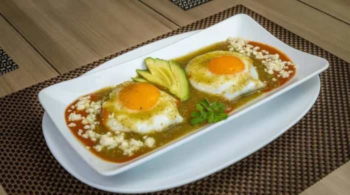 High protein huevos rancheros with two sunny-side-up eggs smothered in red and green salsa, topped with crumbled cotija cheese and avocado slices on a white rectangular plate.