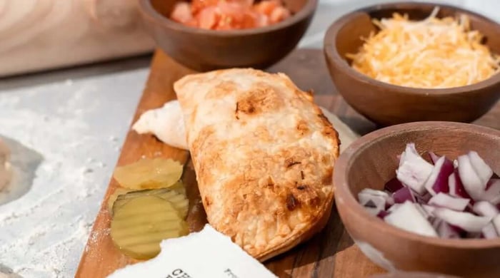 High-protein meal ingredients staged on a wooden board with a baked hand pie, pickles, diced onion, tomato, and shredded cheese.