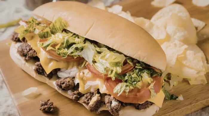 High protein meal featuring a loaded cheesesteak sub with seasoned ground beef, melted cheese sauce, sliced tomatoes, shredded lettuce, and diced onions on a soft hoagie roll.