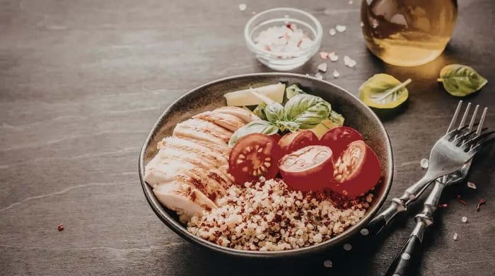 High protein meals featuring sliced grilled chicken breast over quinoa with halved tomatoes, avocado, and fresh basil in a dark ceramic bowl.