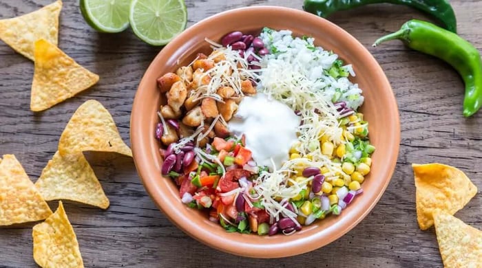 High protein meals featuring a chicken burrito bowl with seasoned chicken, kidney beans, corn, pico de gallo, cilantro rice, sour cream, and shredded cheese in a clay bowl.