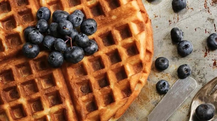 High protein meals with a close-up of a crispy golden round waffle topped with a pile of fresh blueberries on a rustic surface.