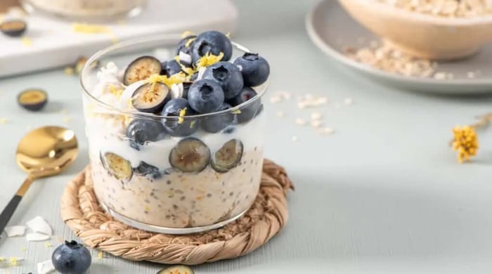 High protein meals with layered overnight oats, chia seeds, yogurt, sliced figs, fresh blueberries, and lemon zest in a clear glass jar on a woven coaster.
