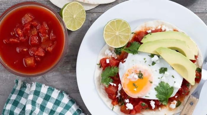 Huevos rancheros served on a flour tortilla topped with red salsa, a fried egg, avocado slices, cotija cheese, and cilantro with a side of salsa roja.