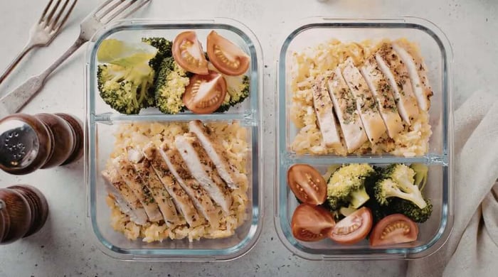 Glass meal prep containers with sliced grilled chicken over yellow rice, broccoli, and cherry tomato wedges from a healthy meal delivery service.