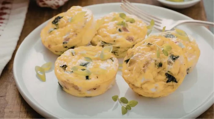 Four fluffy meal prep egg bite cups loaded with spinach, bacon, and cheese topped with fresh oregano on a white plate.