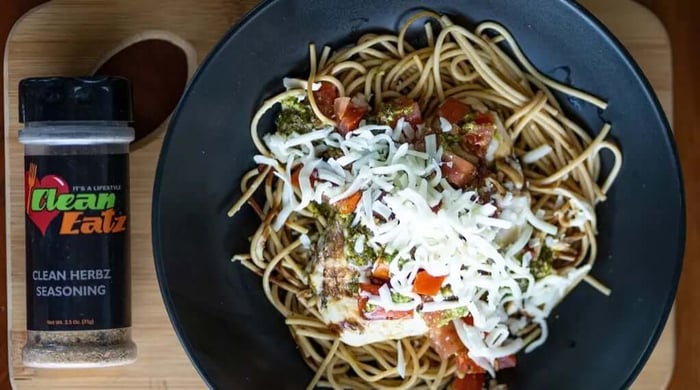 Nutritious ready-made meals with chicken pesto pasta topped with shredded cheese, shown beside Clean Eatz herb seasoning.