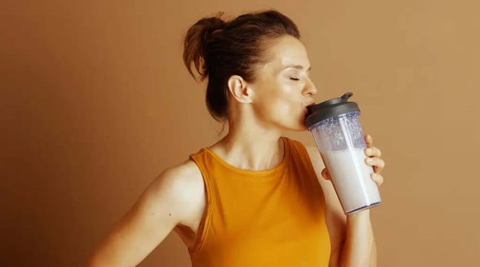 Protein shake for health being sipped by a woman in an orange tank top holding a clear shaker bottle.