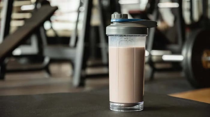 Protein shake in an open shaker bottle sitting on a gym floor surrounded by workout equipment.
