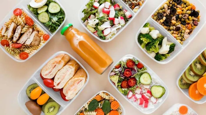 Healthy meal prep spread of bento containers filled with chicken pasta, salads, sandwiches, fruit, and a fresh orange juice bottle.