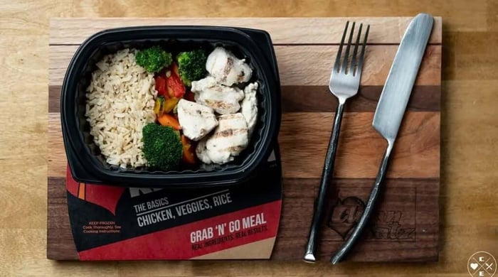 Clean Eatz Kitchen ready-to-eat meal of grilled chicken, broccoli, peppers, and brown rice plated next to a fork and knife on a wood board.
