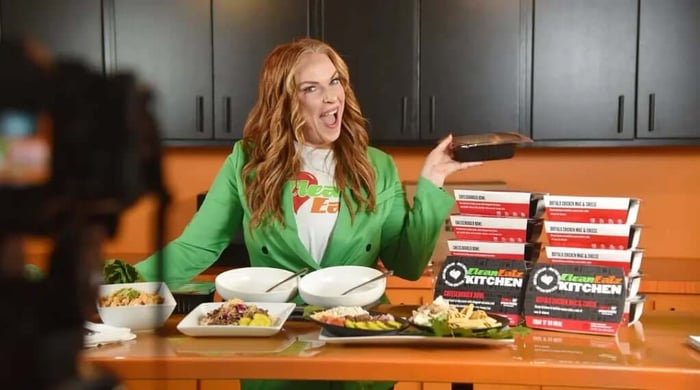 Clean Eatz Kitchen ready-to-eat meals presented on camera by a smiling host in a green blazer holding a stack of branded meal containers.