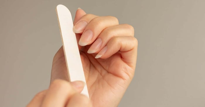 A close-up view shows a person with long, clean nails using a white file to shape the nails against a gray background.