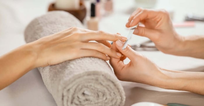 A close-up view shows a person resting their wrist on a rolled towel while someone uses a file to shape their nails.