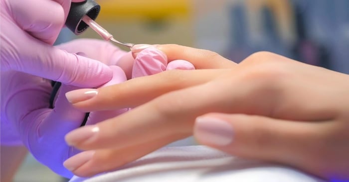 A professional manicurist is painting someone's nails a light pink color. The manicurist is wearing blue gloves.
