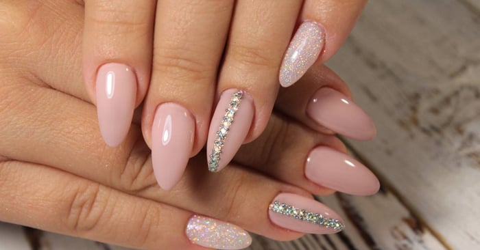 A close-up of almond-shaped nails that are neutral colored with shimmer on the pinky and a strip of rhinestones on the index.