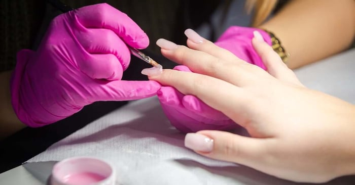 A nail artist is wearing purple gloves and applying a natural pink gel polish to the client's acrylic nail.