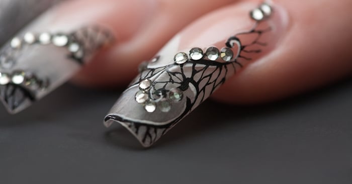 A close-up of a long acrylic nail on someone's hand. The nail features black fine-line designs and silver rhinestones.