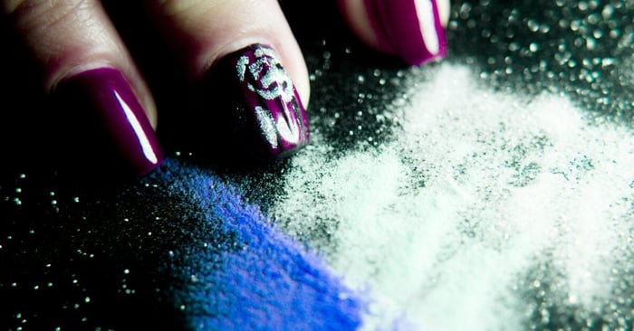 Purple and silver acrylic nails rest on a black, sparkly table, covered in blue and white acrylic powder.