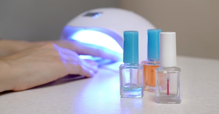 A hand sits in a UV lamp and transparent nail polish bottles stand ready against a white blurred background.