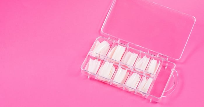 A tidy case of acrylic nail extensions available in different widths and curvatures. The case sits on a pink background.