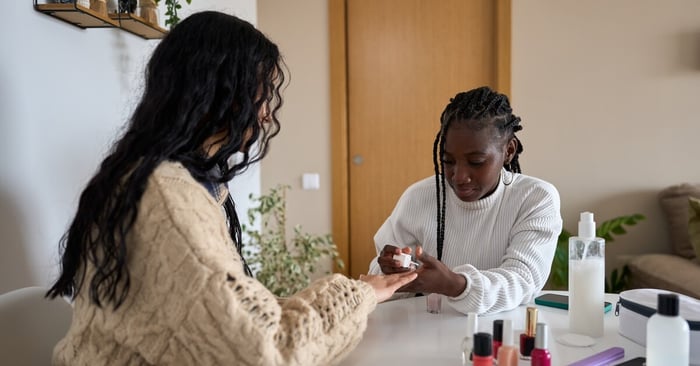 A Black beautician paints a customer's nails at home. Her nail supplies are next to her on a white table.