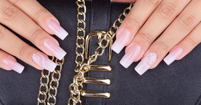 A pair of hands with ombre acrylic nails holds a small black clutch with gold buckles and a gold chain.