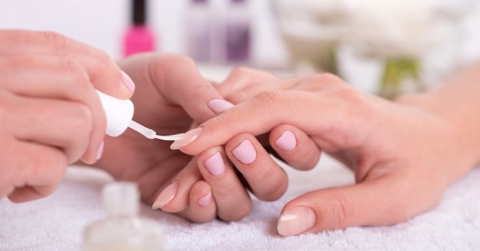 A manicurist uses a nail brush to paint a client’s nails in a professional salon setting with polish bottles in the background.