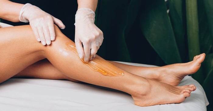 A professional performs a leg waxing treatment on a client's leg in a spa setting, applying warm wax with gloved hands.