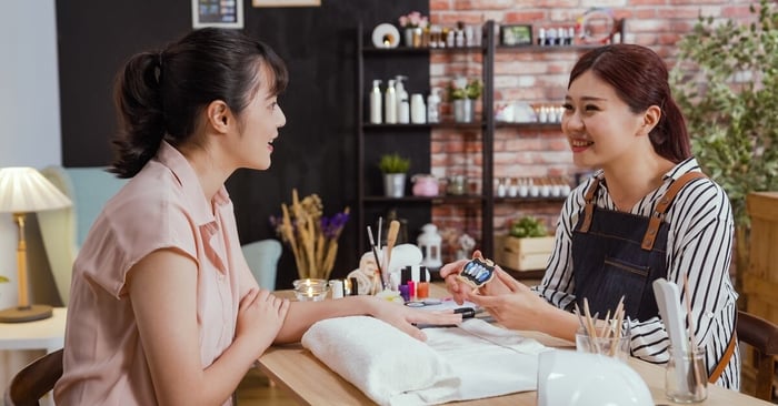 A happy customer sitting across from a nail technician as the technician shows off a small sample of display nails.