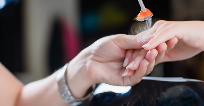 A nail technician holding the hand of a client and using a brush to drop silver nail powder over the nail surface.