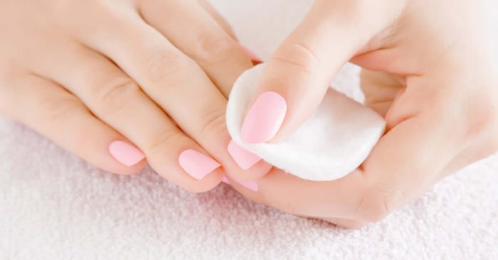 A close-up view shows a person using a white cotton pad to wipe the nail of their index finger, while the hand rests on a towel.
