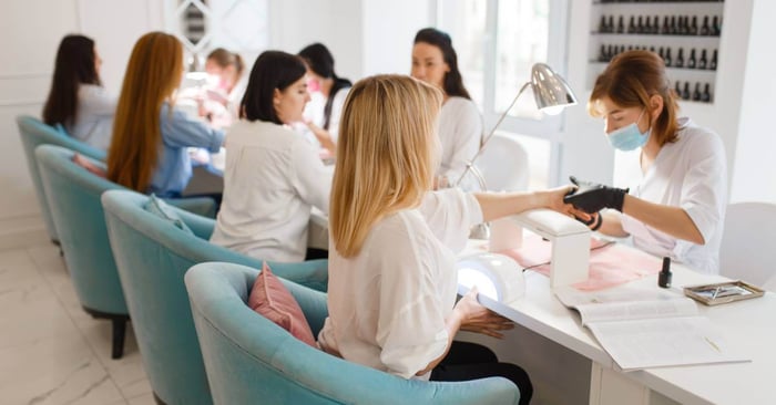 Inside of a nail salon, four women sit in blue chairs at nail stations as they receive manicures from nail technicians.