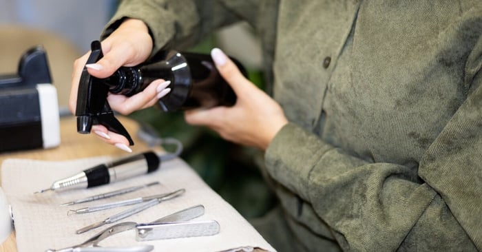 Two hands hold a black spray bottle over various manicure tools, including nail cutters and cuticle pushers, on a desk.