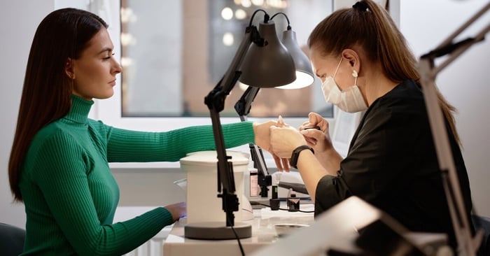 A woman with long, dark hair wearing a green turtleneck sits at a nail salon station and holds a hand out to the technician.