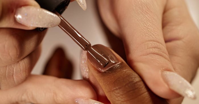 A hand with fair skin holds the hand of a Black client and paints the nails chocolate-brown for a beautiful manicure.