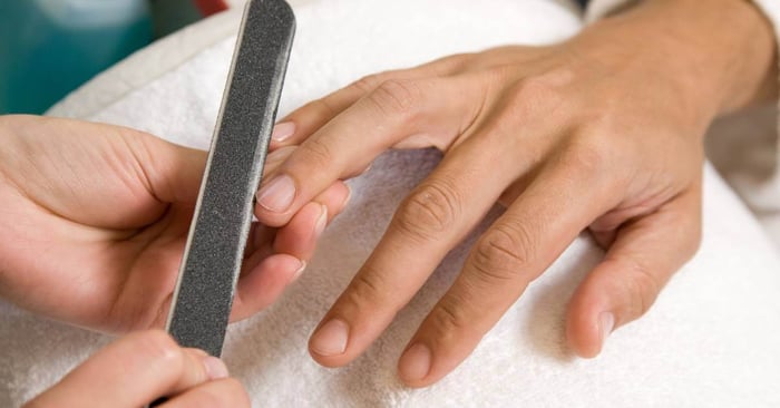 A pair of hands holds the finger of another person’s hand and uses a black nail file to shape the nail.