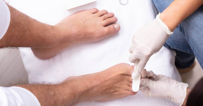 A set of feet placed on top of a white towel as gloved hands from another person position the file to shape the toenails.