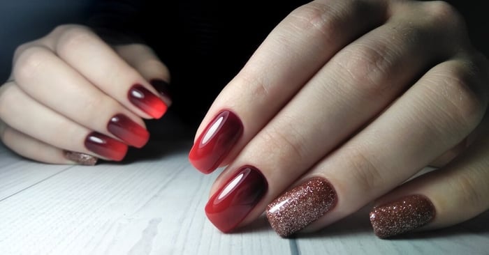 A close-up of someone's burgundy-cherry red gradient nails. The ring and pinky fingernails have glitter polish on top.
