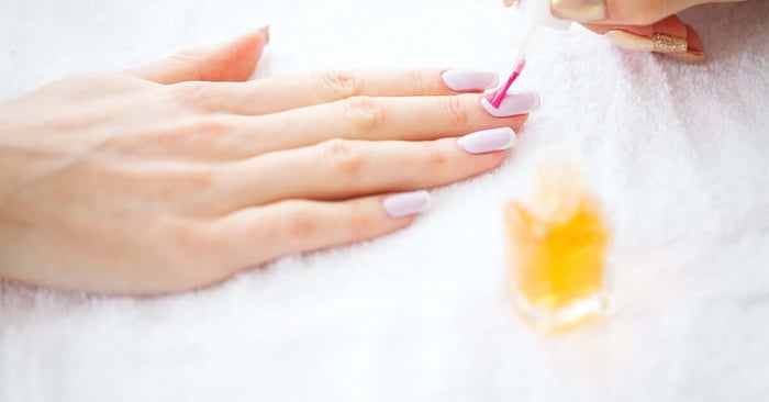 A close-up of a woman's manicured hand resting on a white towel. A nail tech is putting oil on her nails.