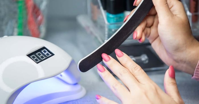 A person with long pink nails files their manicure with a nail shaper. An ultraviolet lamp is near their hands.
