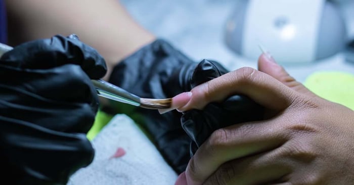 A close-up of the process of applying acrylic powder on the nails. The nail tech is wearing black gloves.